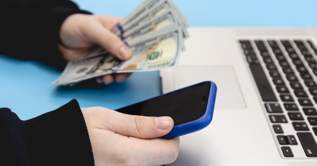 Person holding cash and using a smartphone beside a laptop