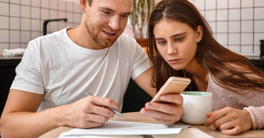 Couple sitting at their kitchen table reviewing mortgage documents and comparing interest rate and APR on a phone