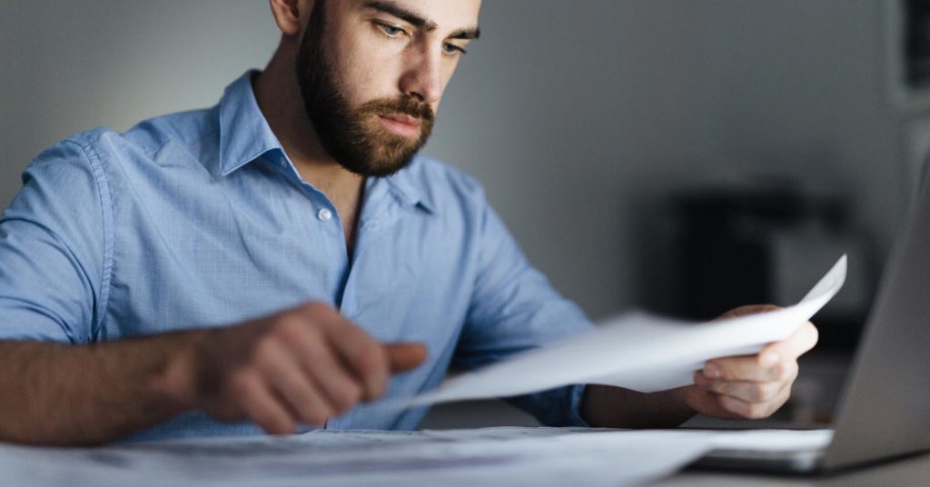 Man comparing interest and fees on debt collection statements beside a laptop