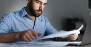 Man comparing interest and fees on debt collection statements beside a laptop