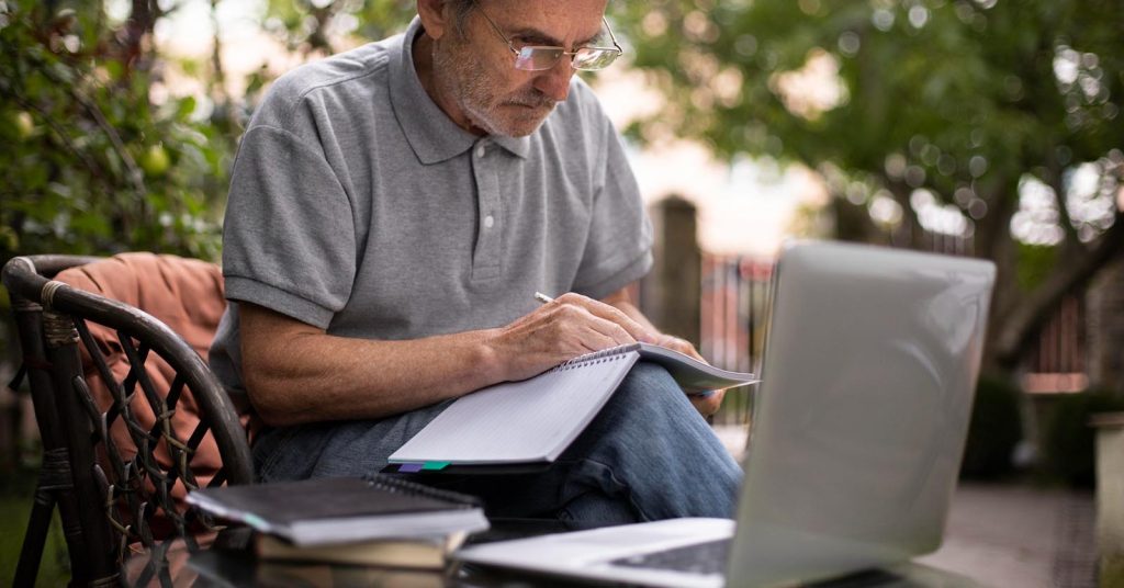 Older man writing in a notebook beside a laptop outdoors