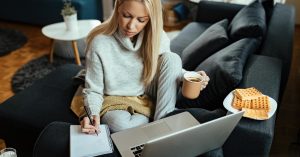 Woman using a laptop and writing notes while sitting on a sofa