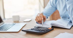 Person using a calculator beside a laptop and open notebook