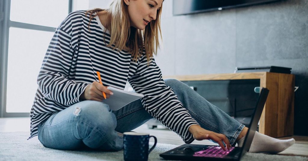 Woman taking notes while using a laptop on the floor