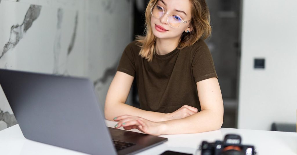 Woman using a laptop at a table with a camera and smartphone nearby