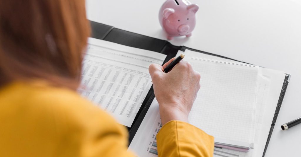 Person writing on a financial worksheet beside a pink piggy bank