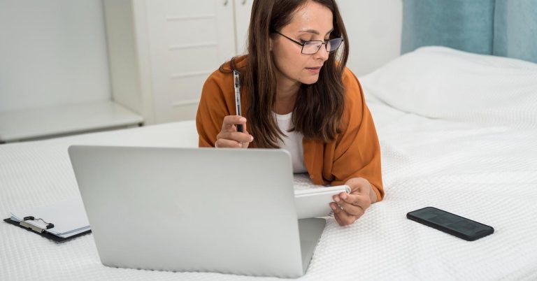 Woman reviewing notes while checking financial information on a laptop