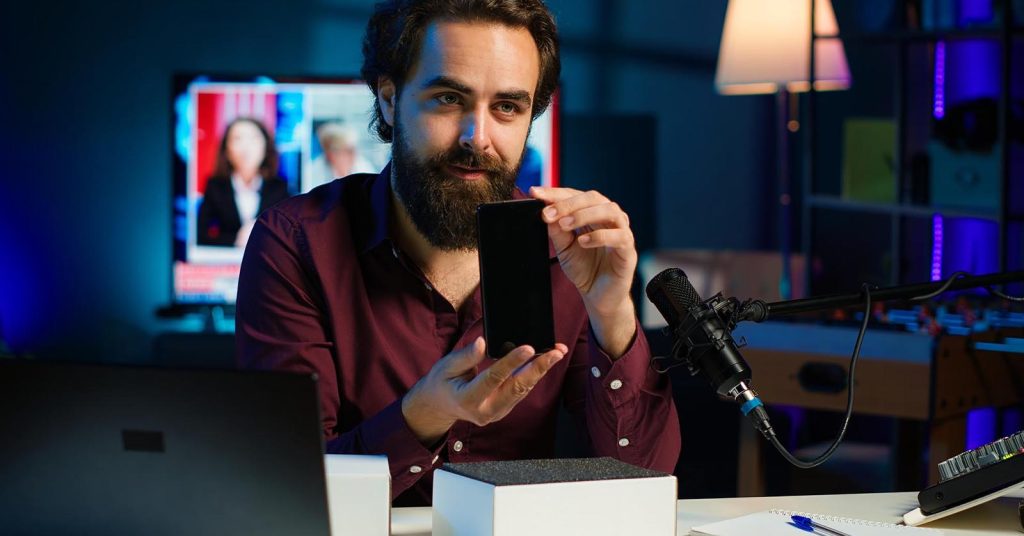 Man presenting a smartphone at a desk with a microphone and laptop