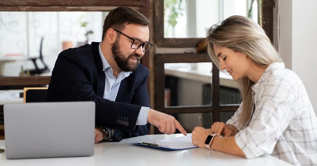 Woman signing a document while another person points to the paperwork