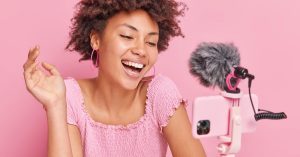 Woman recording a video with a smartphone and external microphone against a pink background
