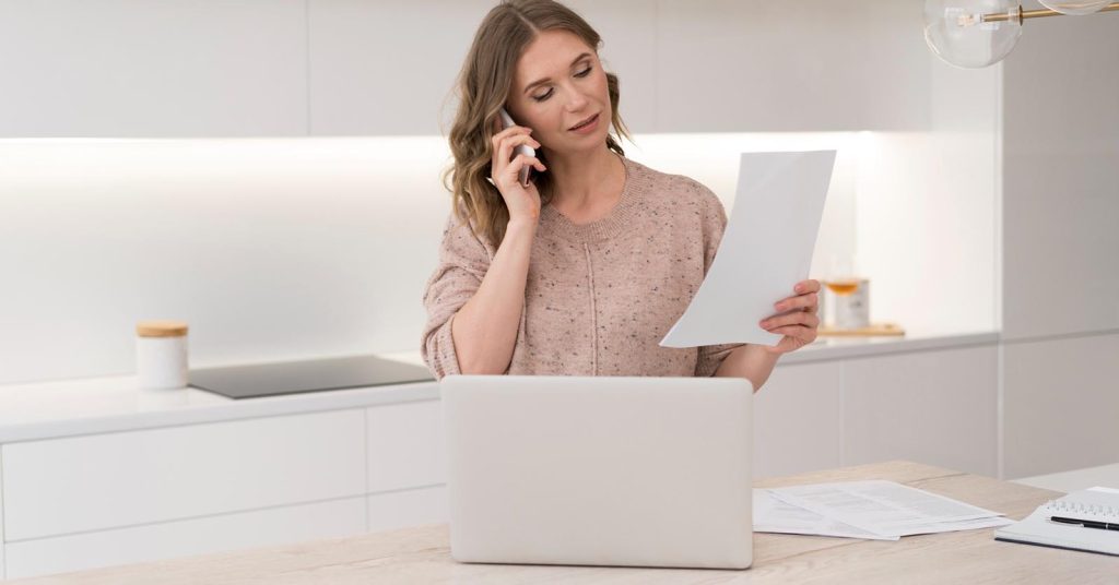 Woman speaking on the phone while holding a document beside a laptop