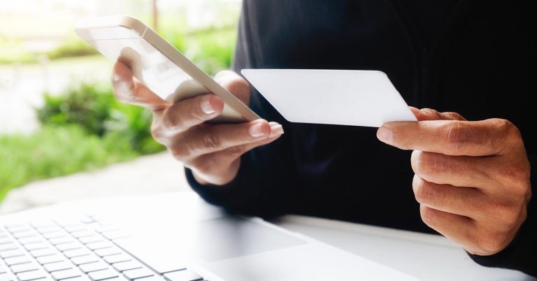 Person holding a credit card and smartphone beside a laptop