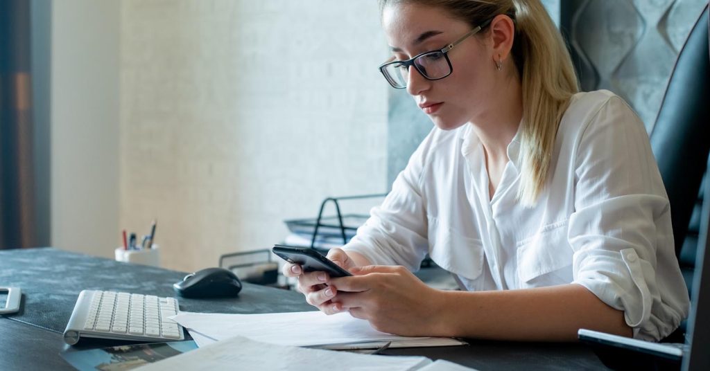 Woman checking a smartphone while sitting at a desk with financial papers