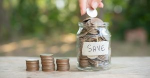 Hand dropping a coin into a savings jar labeled SAVE beside stacks of coins
