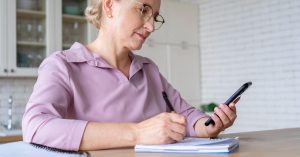 Older woman writing notes while using a smartphone at home