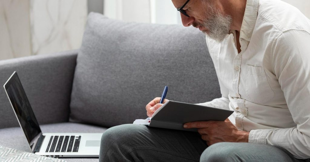 Older man writing in a notebook beside a laptop at home