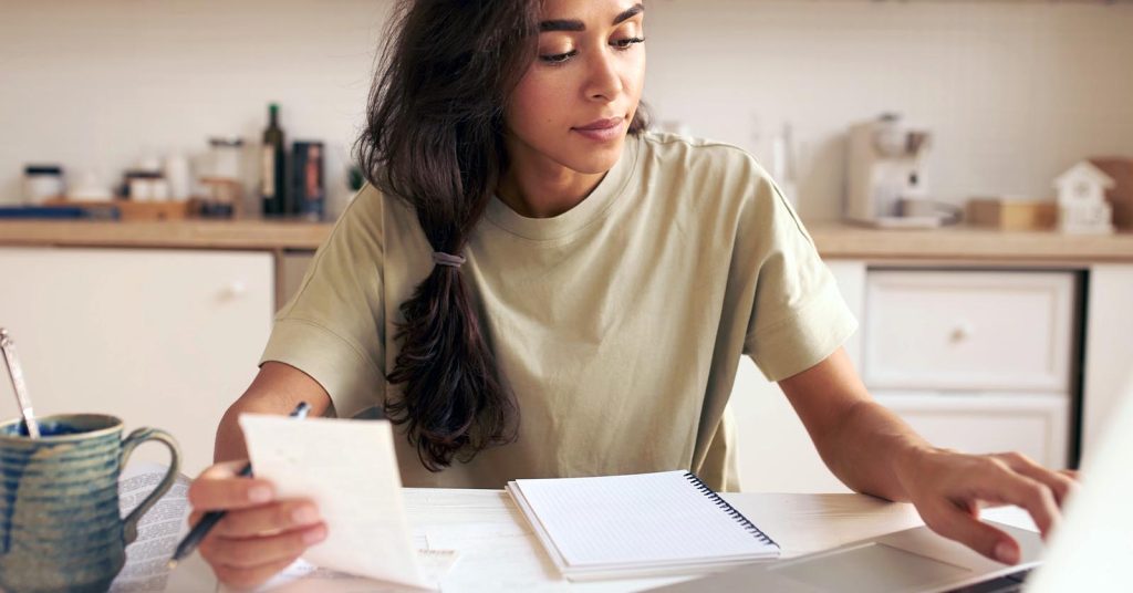 Woman reviewing paperwork and using a laptop at a kitchen table