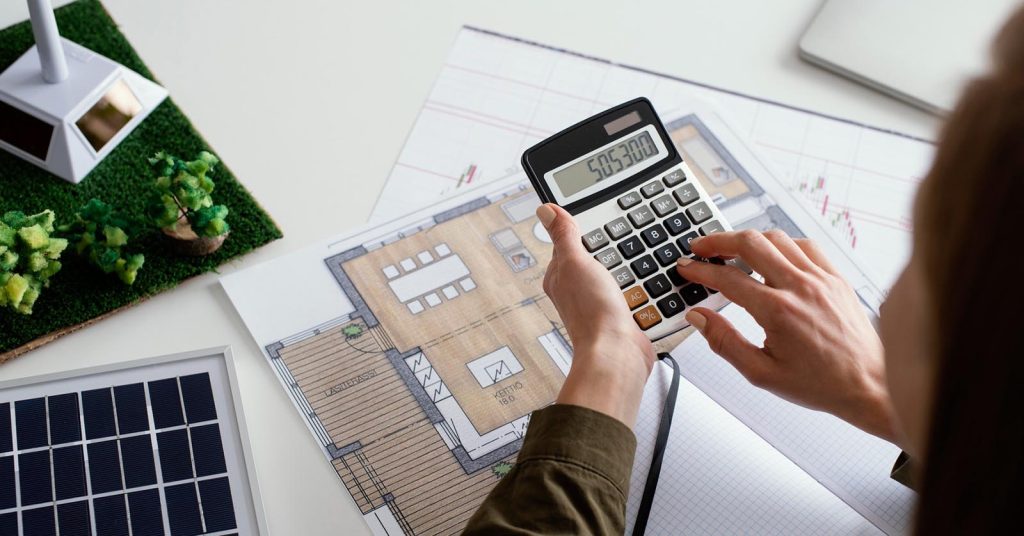 Person calculating home energy costs beside house plans and a solar panel model