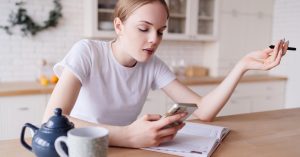 Woman looking at a smartphone while sitting at a table with an open notebook