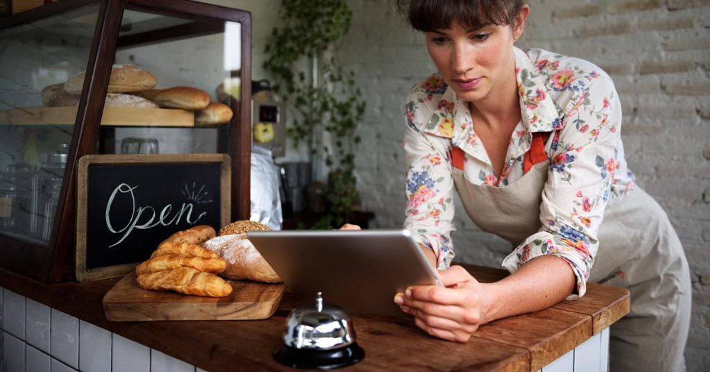 Woman using a tablet behind a bakery counter with an Open sign