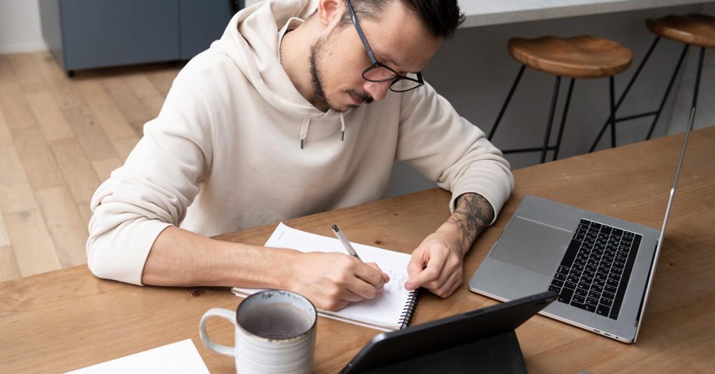 Man consistently tracking debt payoff progress in a notebook beside a laptop and tablet