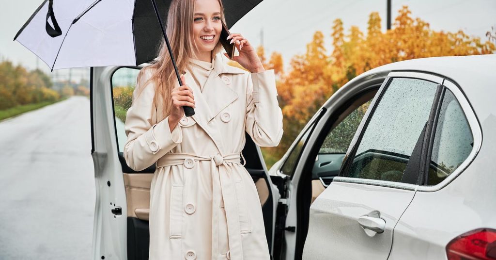 Woman holding an umbrella and talking on the phone beside a white car
