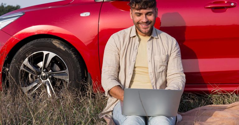 Man using a laptop while sitting beside a red car