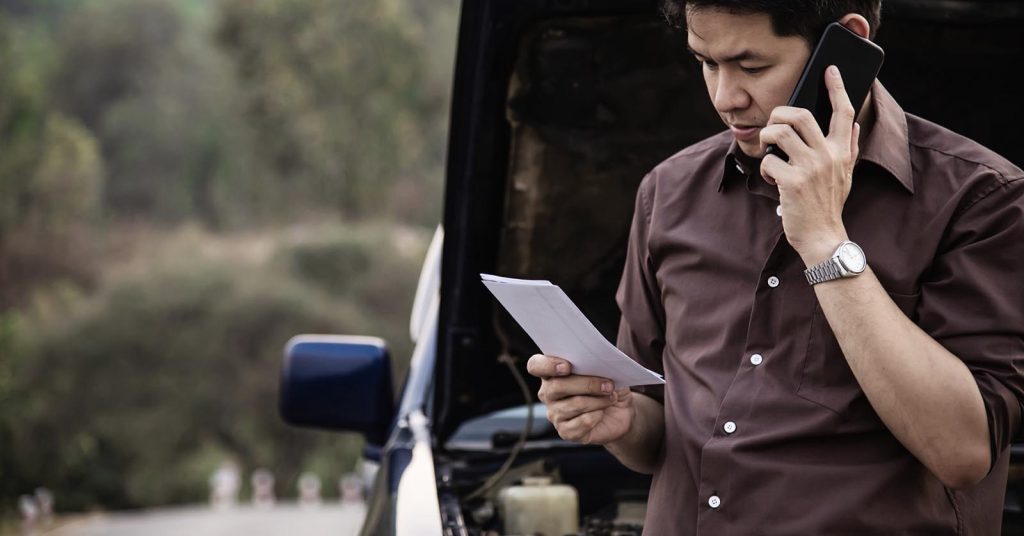 Man speaking on the phone and holding a document beside a car with the hood open