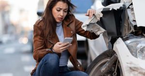 Woman using a smartphone beside a badly damaged car after a crash