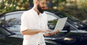 Man using a laptop while standing beside a parked car